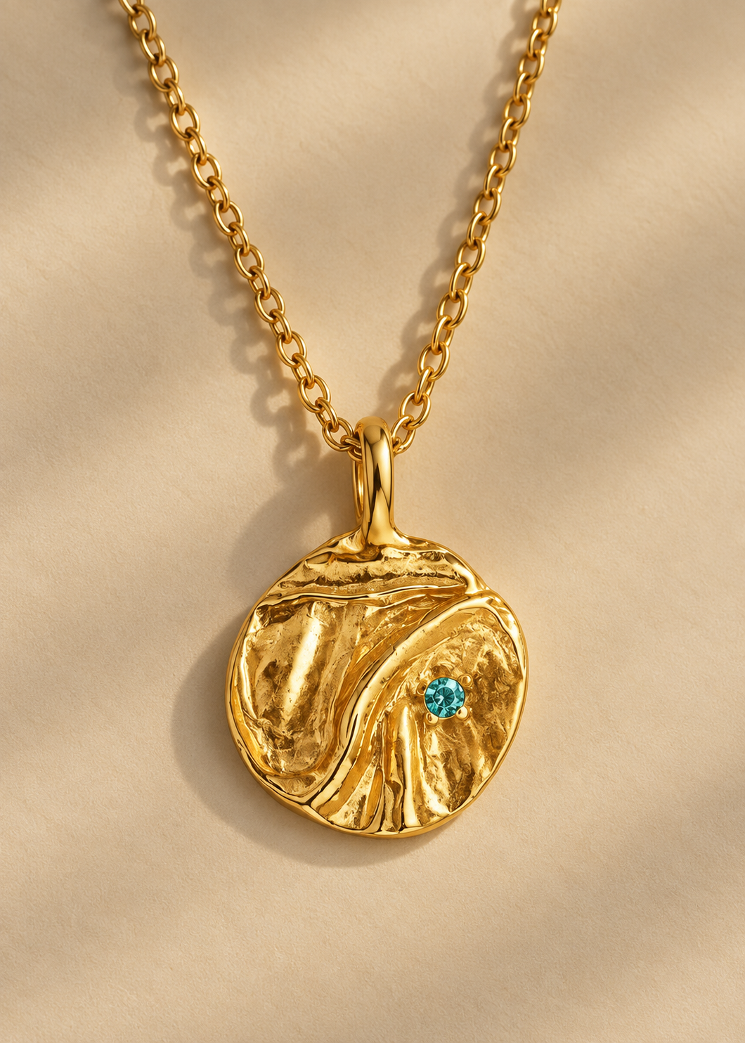 A textured gold pendant necklace with a small turquoise gemstone, displayed on a soft beige fabric background with elegant shadows.