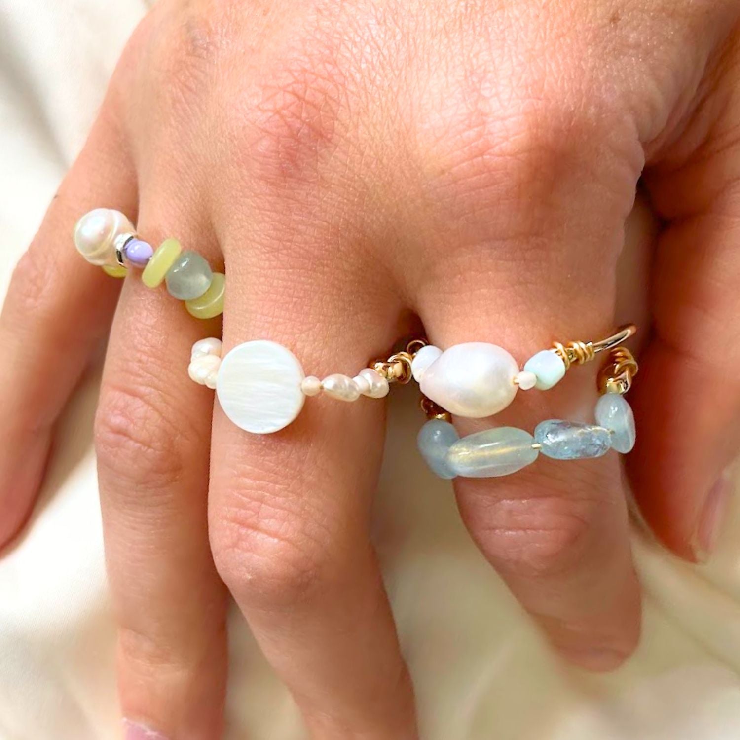 Close-up of a hand wearing two beaded rings with pearls and gemstones on a neutral background.