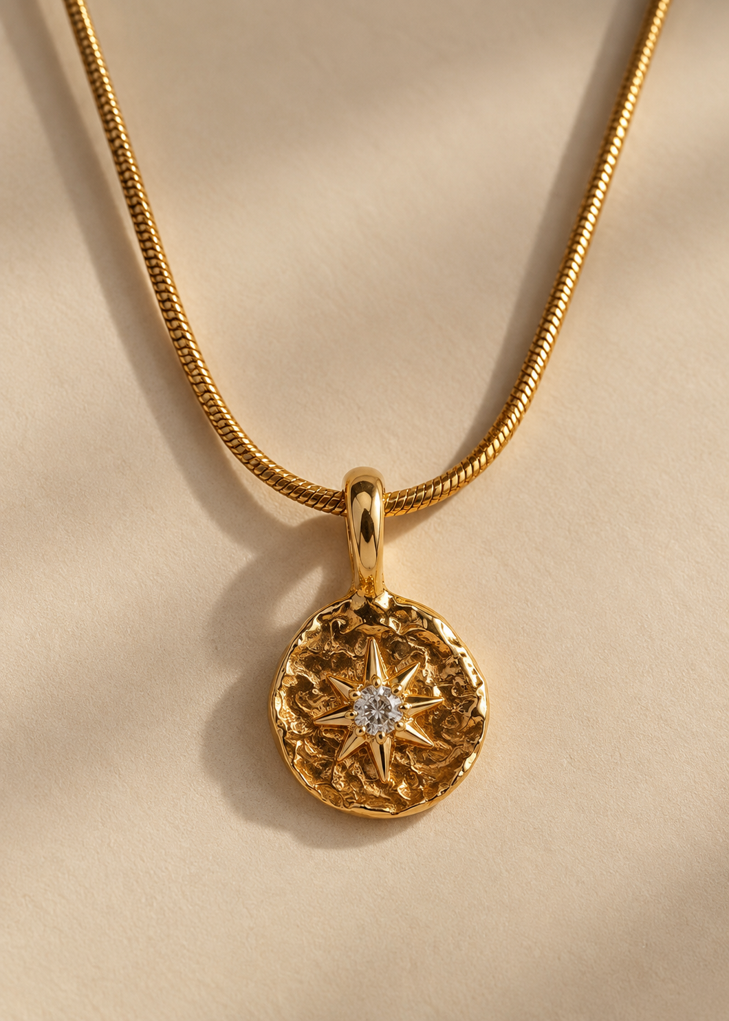 A gold pendant necklace featuring an ornate circular medallion with a starburst motif and a small clear gemstone at the center, suspended from a gold chain on a warm beige background.