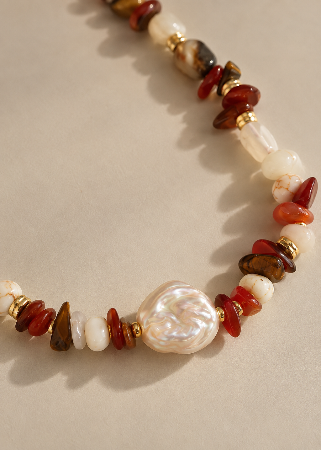 A beaded necklace with white baroque pearls, polished red and brown stone chips, and a central irregular white pearl pendant, displayed on a neutral beige background.