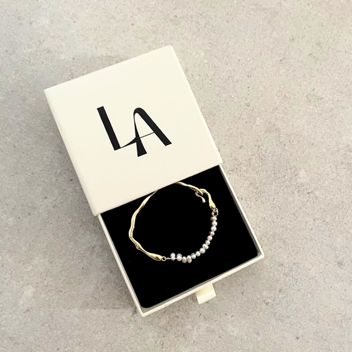 Gold and pearl bracelet in a box with 'LA' initials on a gray surface