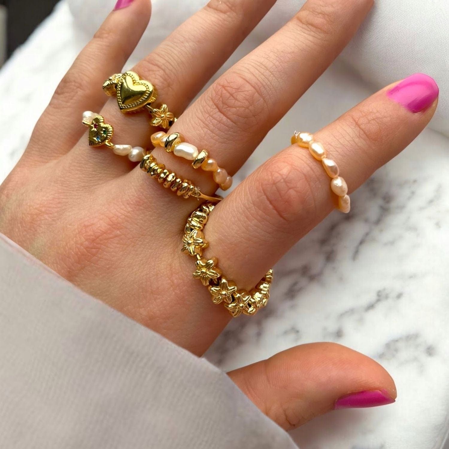 Hand wearing multiple gold and pearl rings with pink nail polish on a marble background