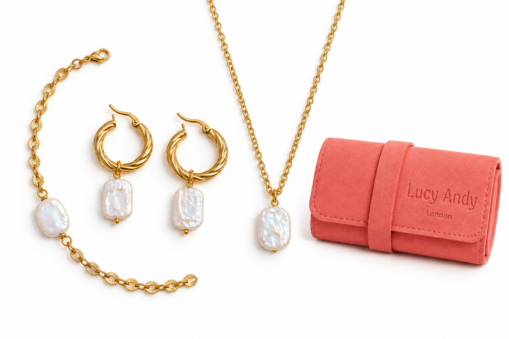 Gold jewelry set with baroque pearl drops, including a necklace, hoop earrings, and bracelet, displayed next to a coral pink faux-leather roll-up case embossed with 'Lucy Andy London' on a white background.