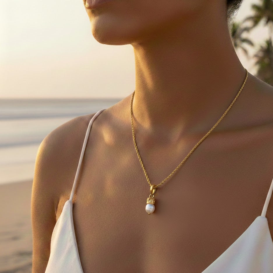Woman wearing a gold necklace with a pearl pendant on a beach.